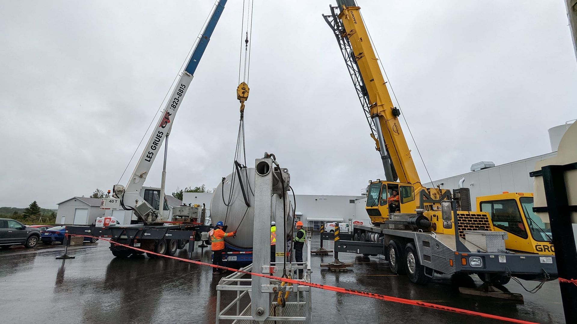 Deux grues soulèvent une grande structure blanche sur un chantier pluvieux
