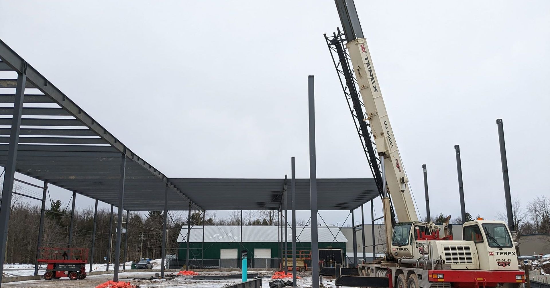 Chantier de construction avec un bâtiment à ossature métallique et une grue soulevant des poutres par temps couvert