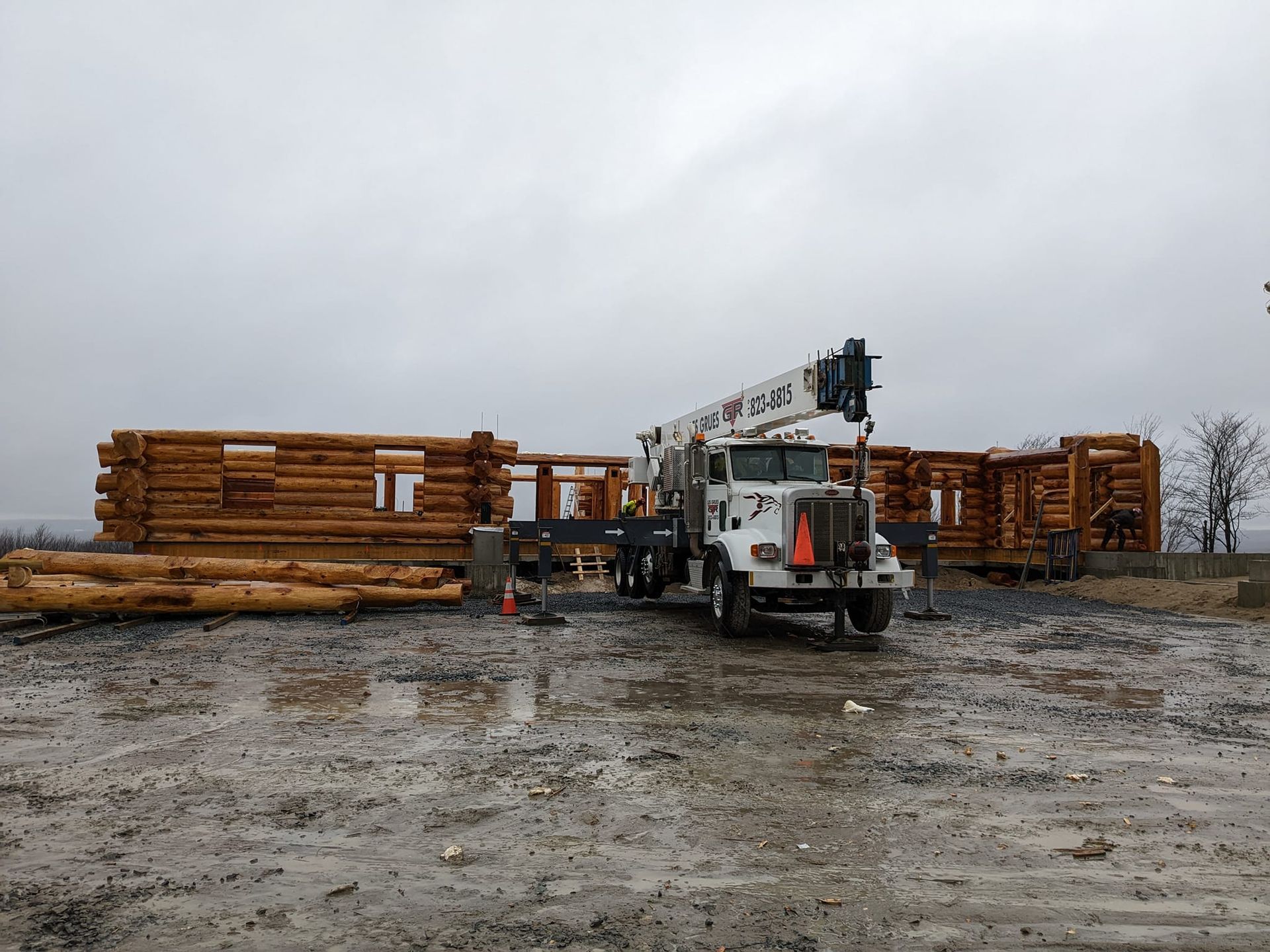 Camion grumier avec grue dans une cour boueuse à côté de bois empilé sous un ciel couvert
