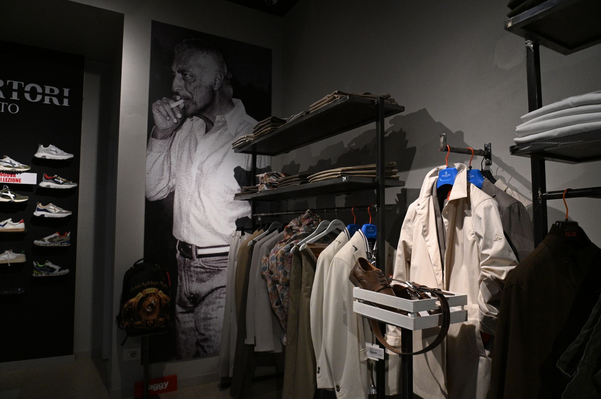 Interno di un negozio di abbigliamento con scaffali pieni di vestiti, scaffalature e un grande poster di un uomo.