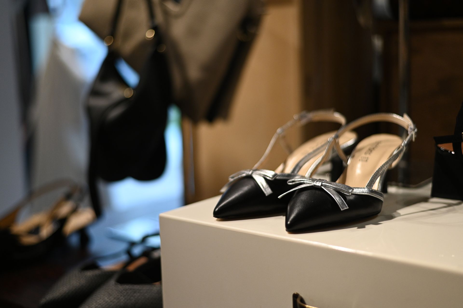 Scarpe con tacco a spillo nere e argento con fiocchi su una superficie bianca, con borse sullo sfondo.