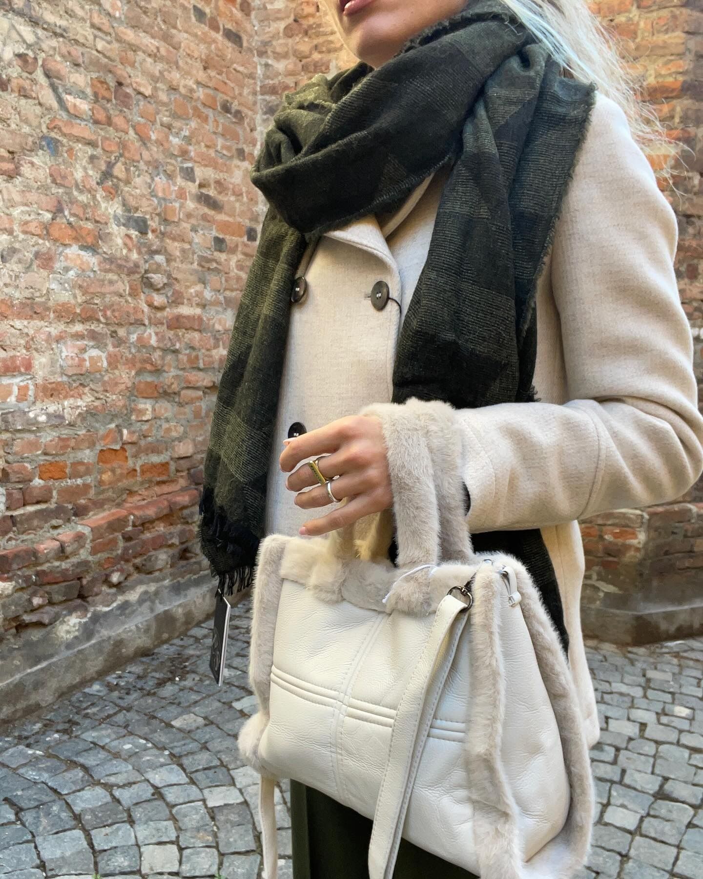 Donna con cappotto color crema e sciarpa verde, che tiene in mano una borsa color crema appoggiata a un muro di mattoni.
