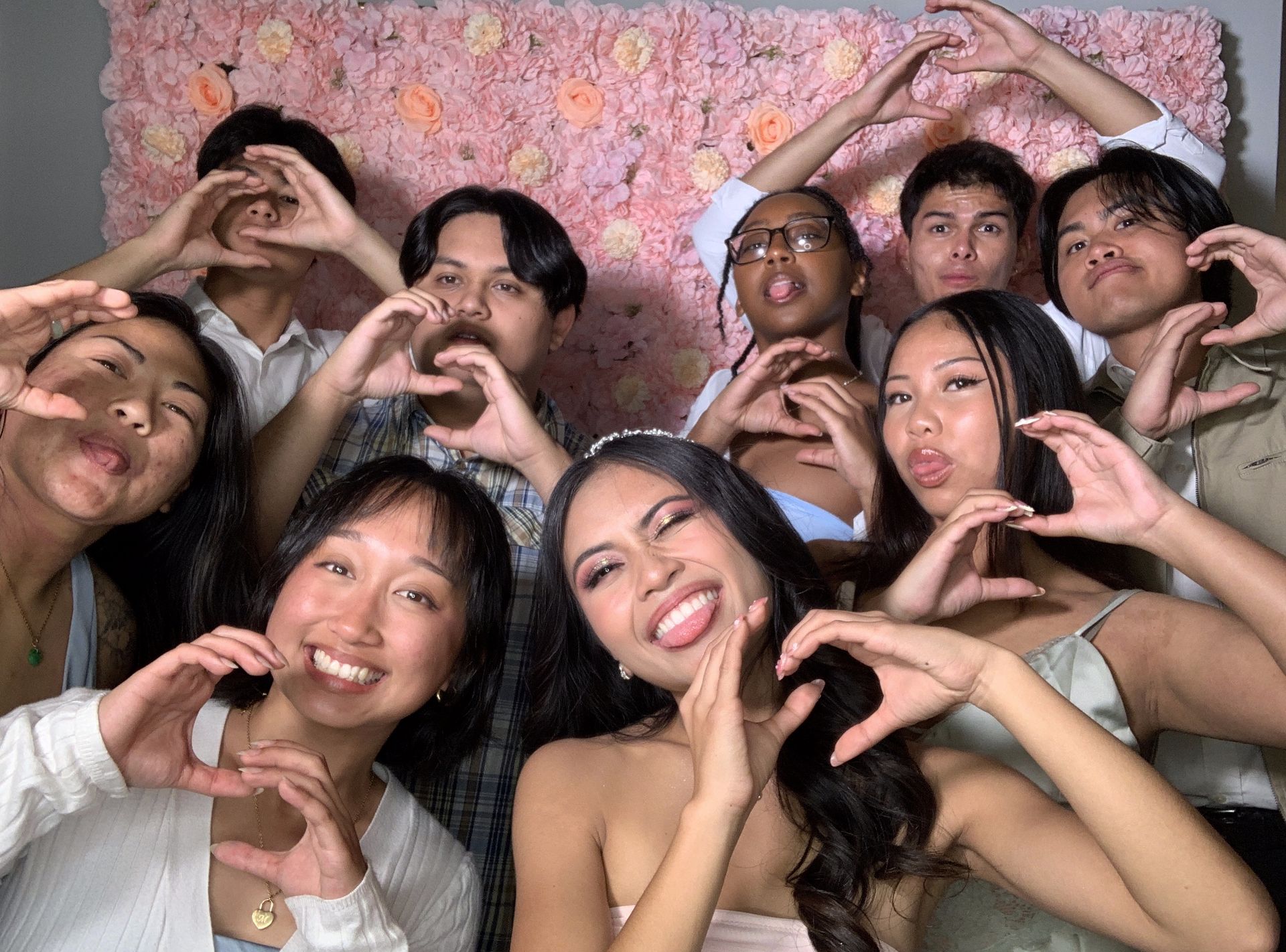 Group of friends posing in front of a pink floral wall, making heart shapes with their hands and smiling.