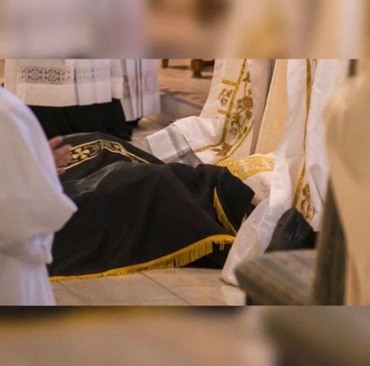 Person in black-and-gold ceremonial robe lying on the floor among white robes indoors