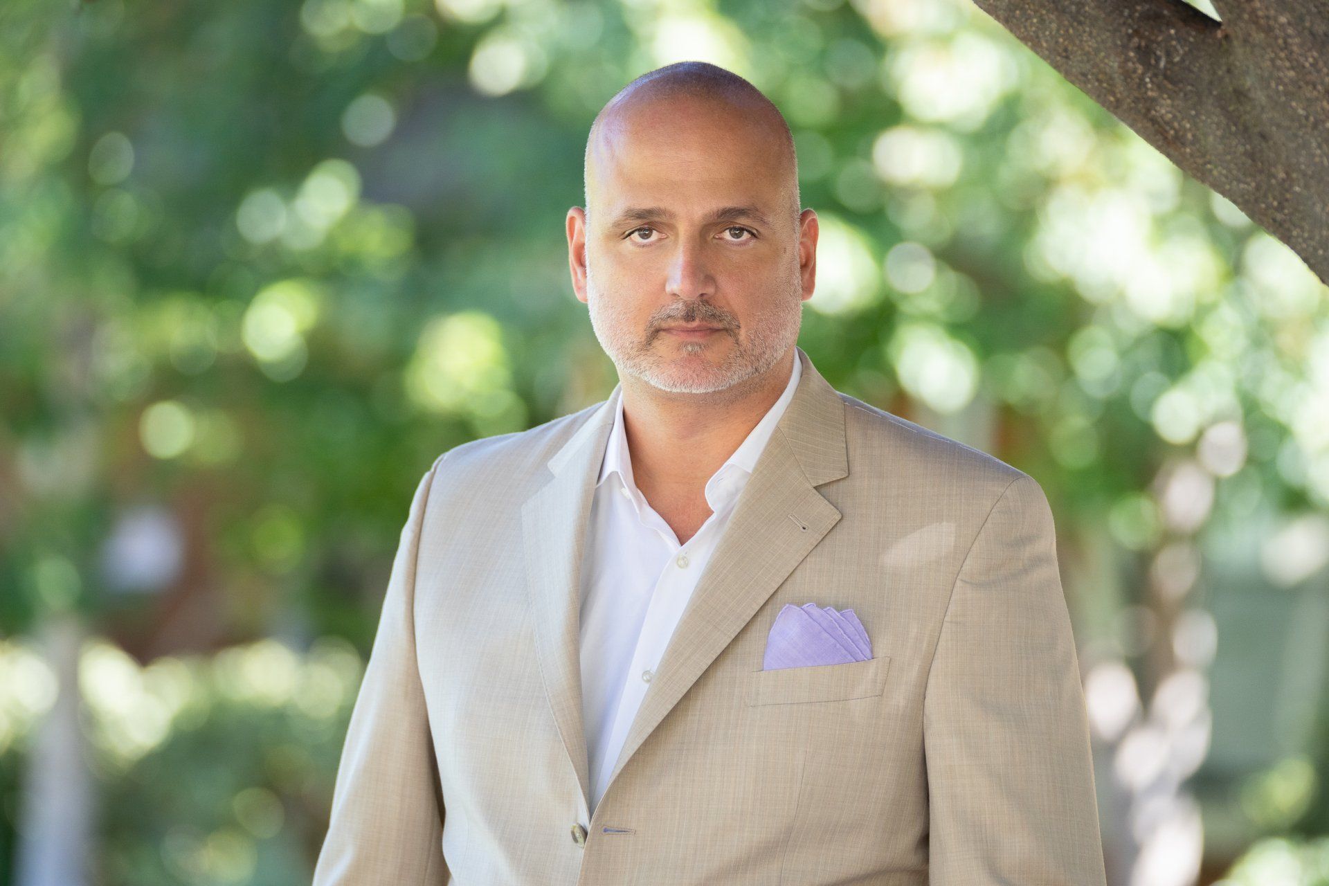 A man in a tan suit and white shirt is standing under a tree.