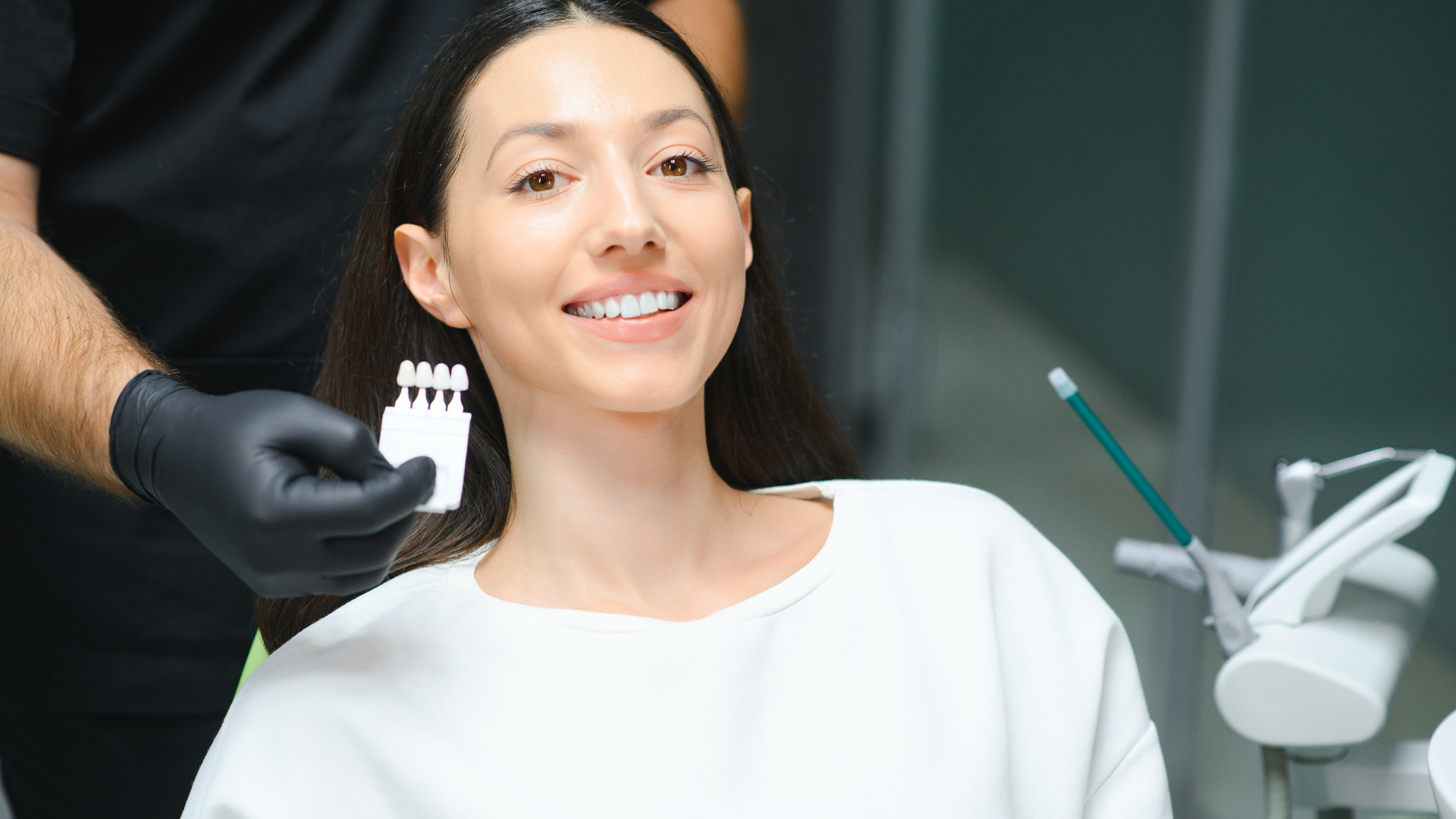Woman smiling at dentist, teeth shades held up for comparison. Dental office setting.
