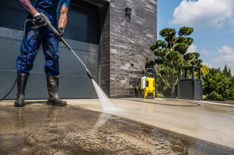 Worker pressure-washes a concrete driveway beside a modern building on a sunny day