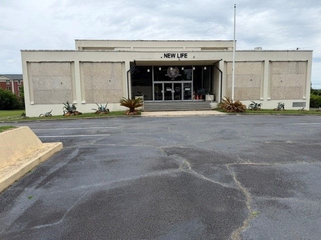Front entrance of a beige building with a paved parking lot in front, cloudy sky overhead