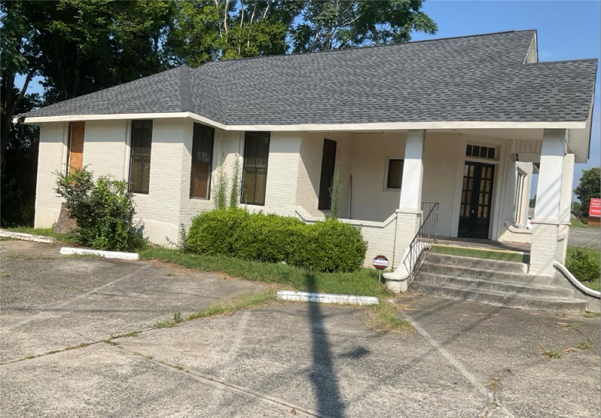 Single-story beige house with white columns, front steps, and shrubs in a sunny driveway.