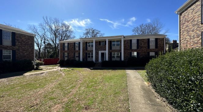 Brick apartment building with white columns, front lawn, and walkway under a clear blue sky