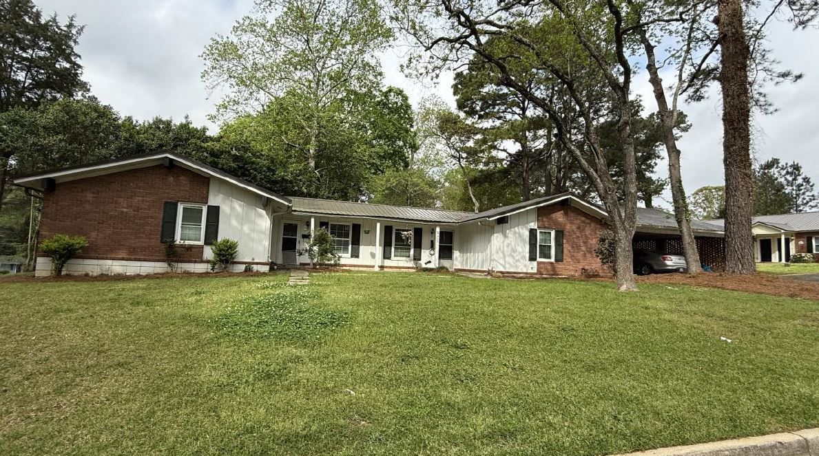 Single-story suburban house with brick and white siding, set behind a large grassy yard and tall trees.