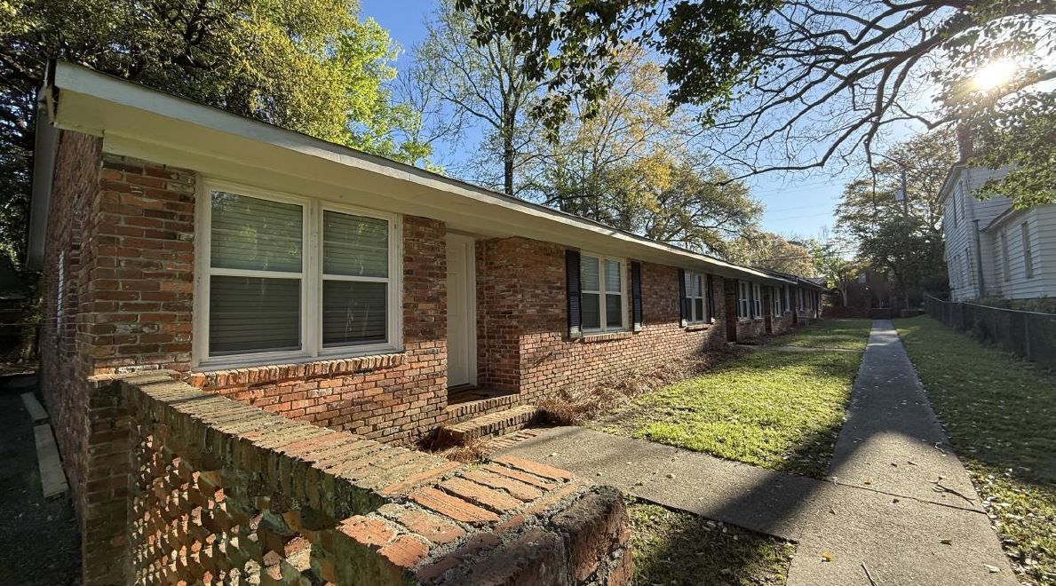 Brick apartment building with white shutters and a sidewalk beside a grassy courtyard in sunlight