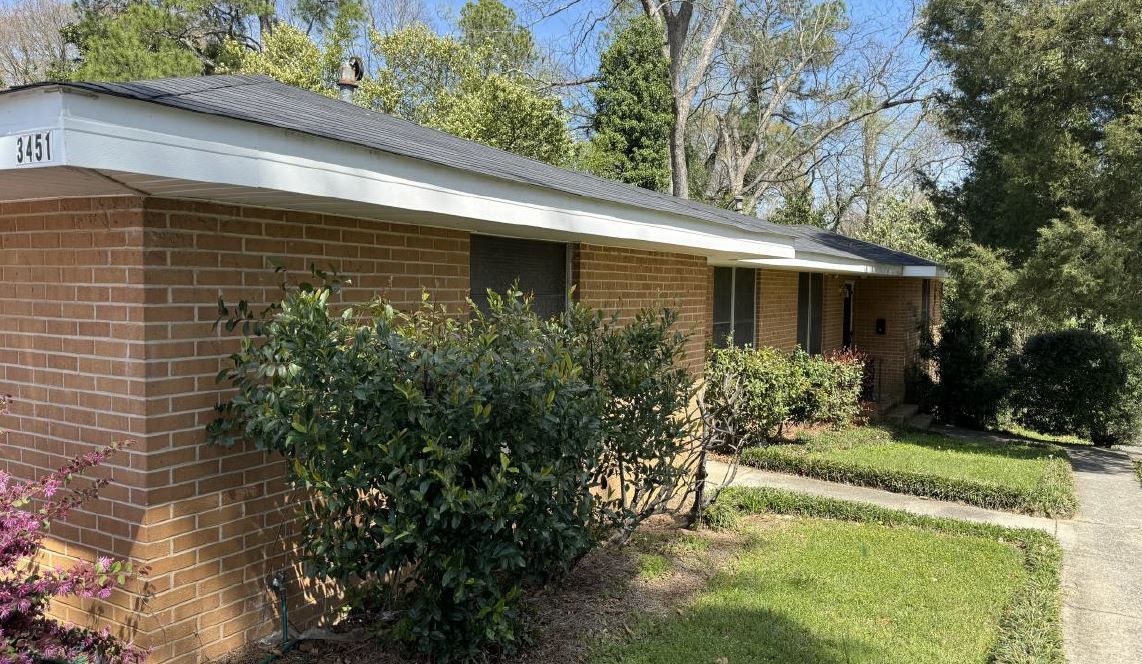 Single-story brick house with white trim, bushes, and a concrete walkway under a sunny sky