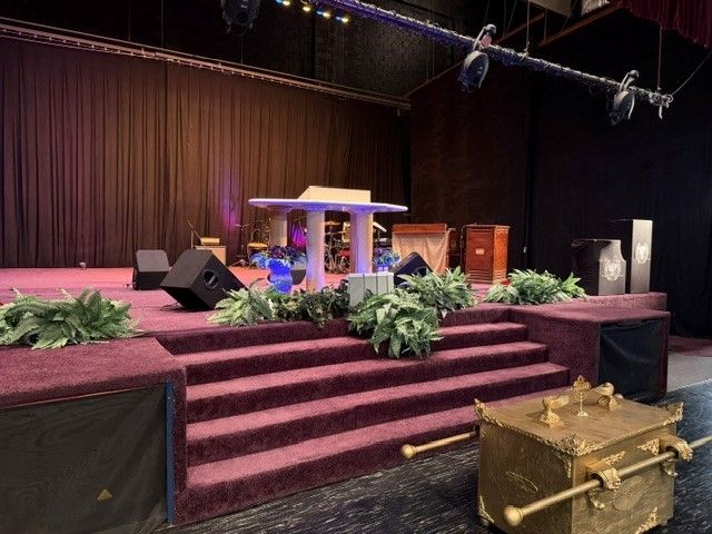 Indoor stage with purple carpeted steps, lectern, plants, and draped curtains under stage lighting