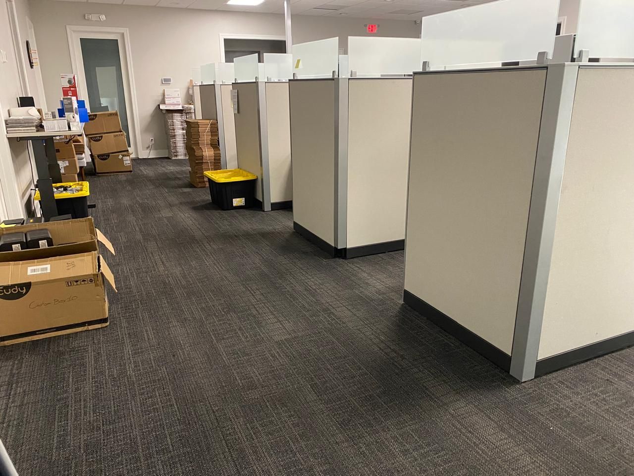 Office hallway with beige cubicle partitions, carpeted floor, and stacked boxes along the left wall