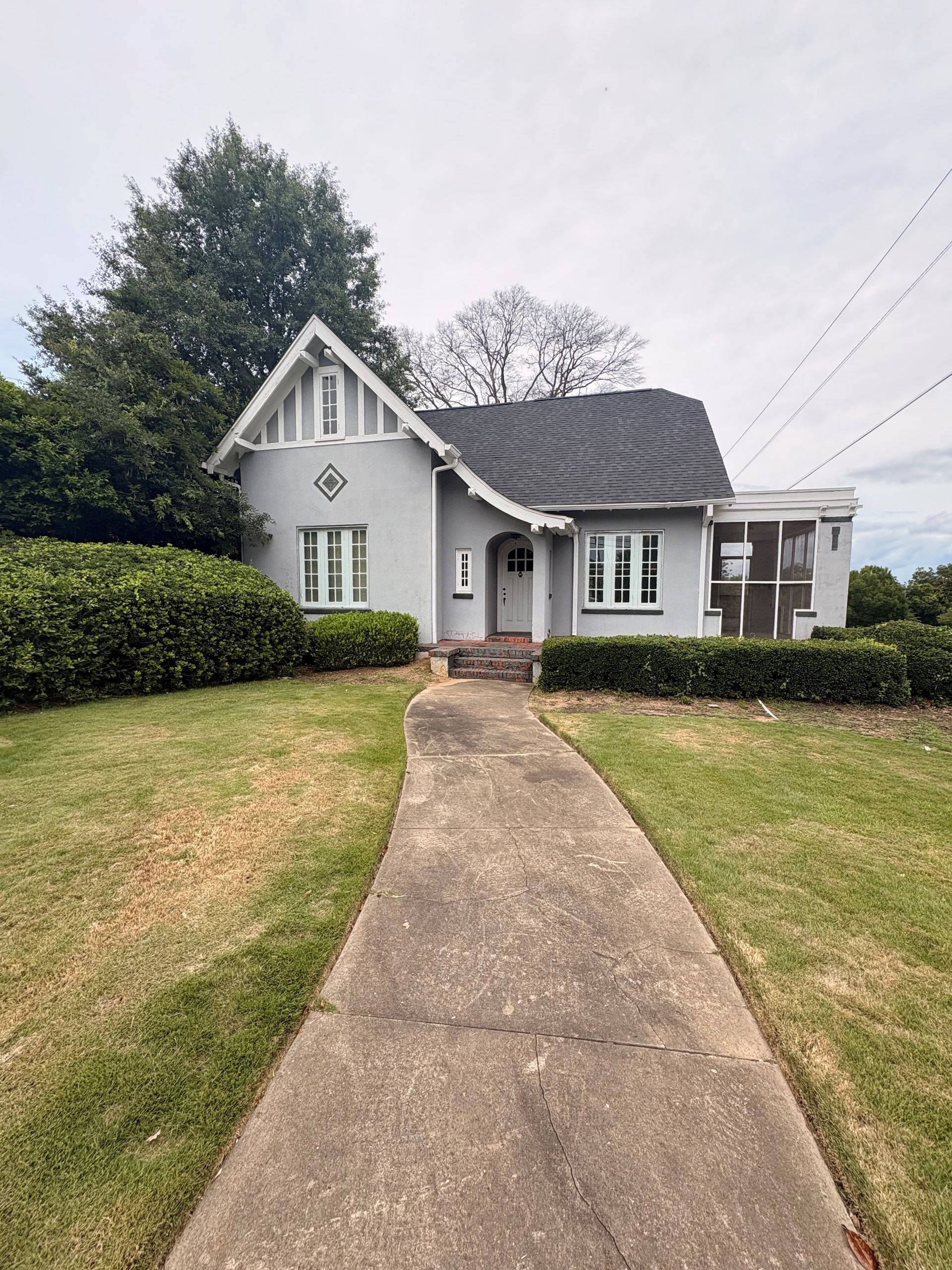 Gray cottage with a steep roof, front walkway, trimmed hedges, and a grassy yard.