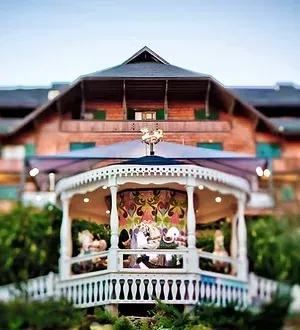 Gazebo branco com fundo floral em frente a uma construção rústica de madeira e um jardim verdejante.