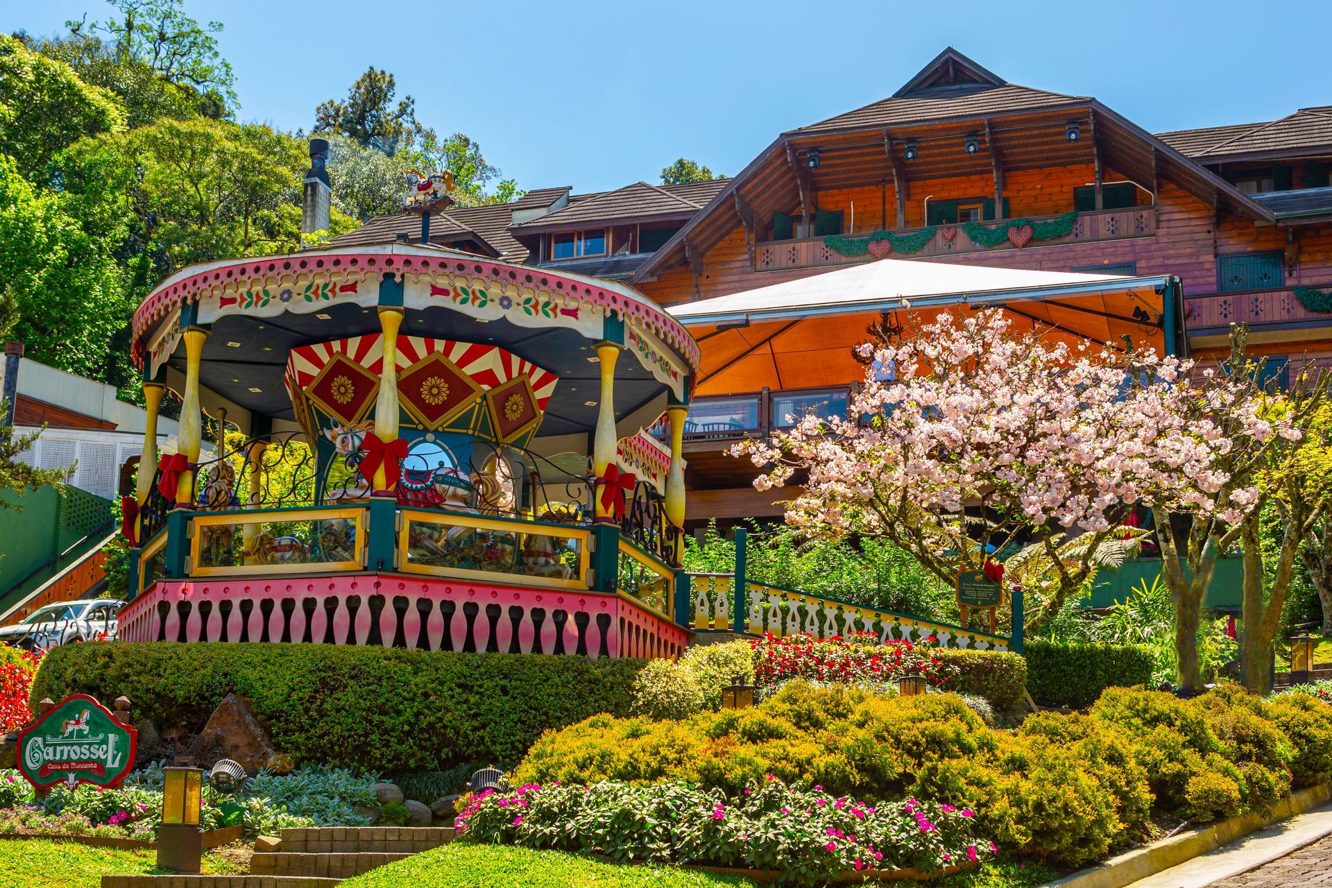 Gazebo branco com fundo floral em frente a uma construção rústica de madeira e um jardim verdejante.