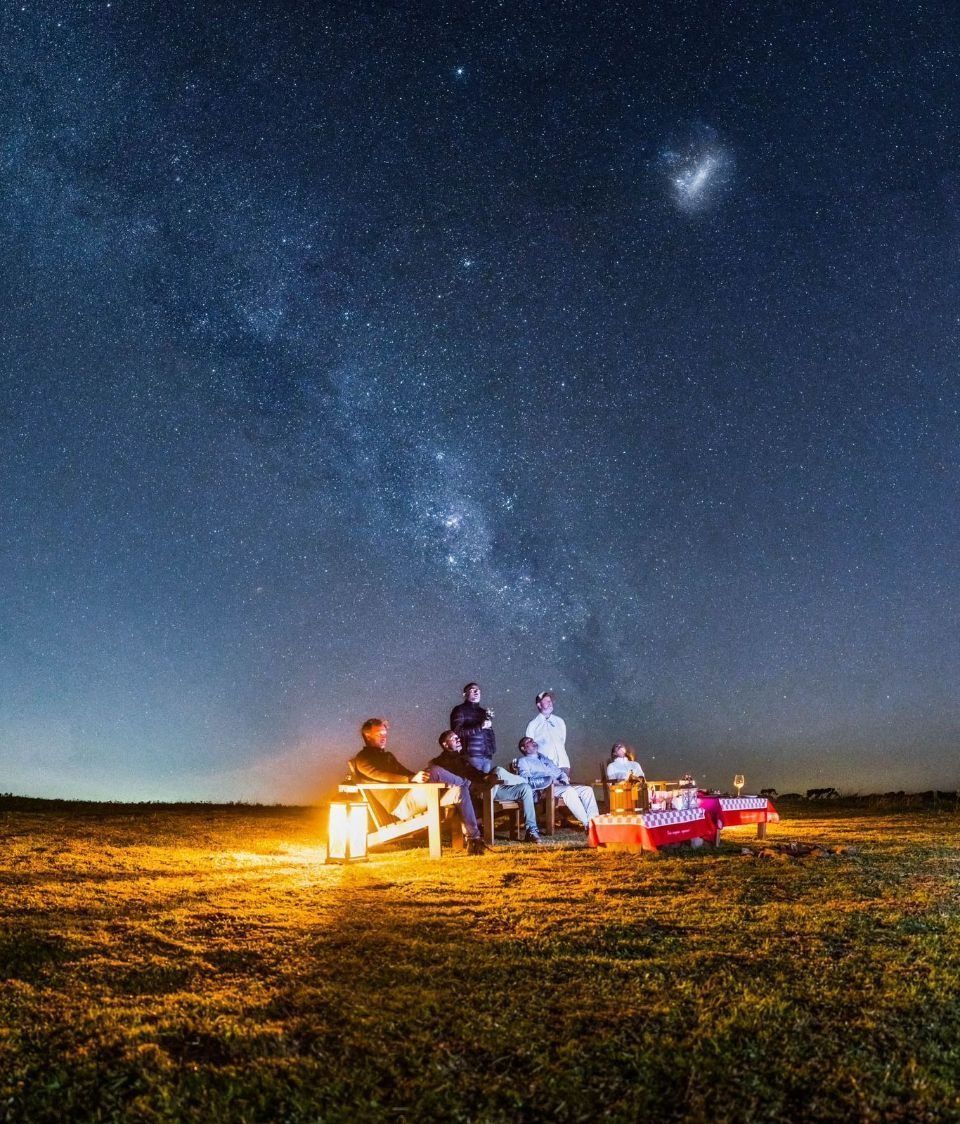 Pessoas sentadas ao redor de uma fogueira sob um céu estrelado em um campo aberto.