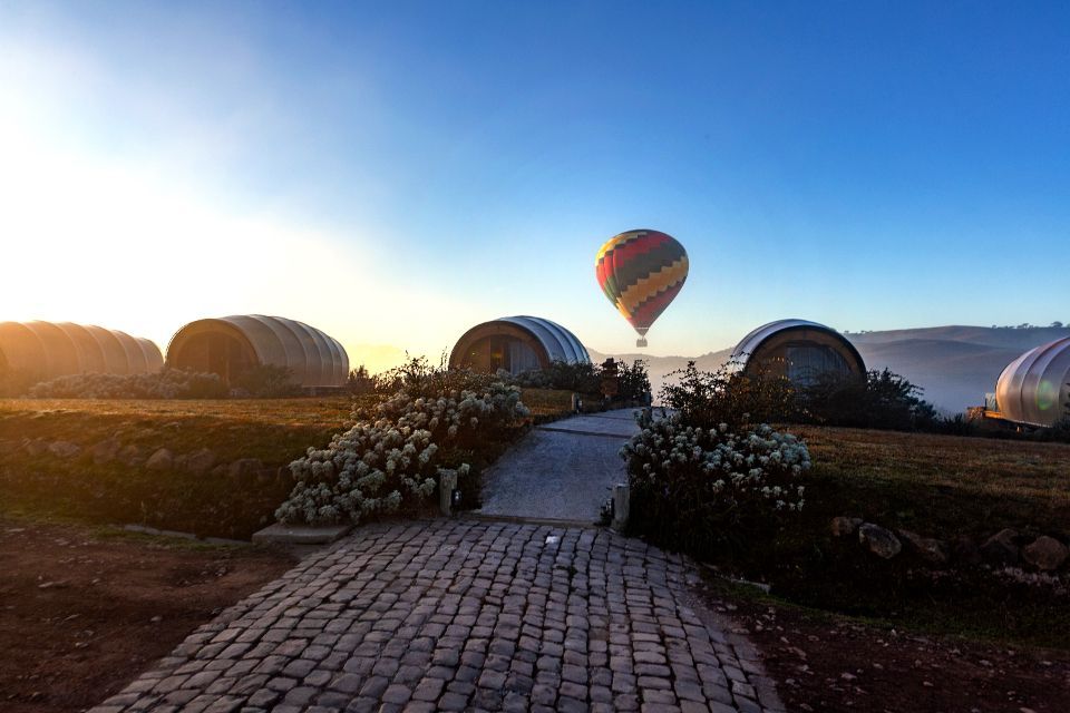 Nascer do sol sobre um caminho de paralelepípedos que leva a tendas em forma de cúpula, com um balão de ar quente em um céu azul claro.
