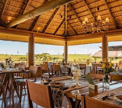 Restaurante ao ar livre com mesas postas sob um telhado de palha, com vista para uma paisagem ensolarada de savana.