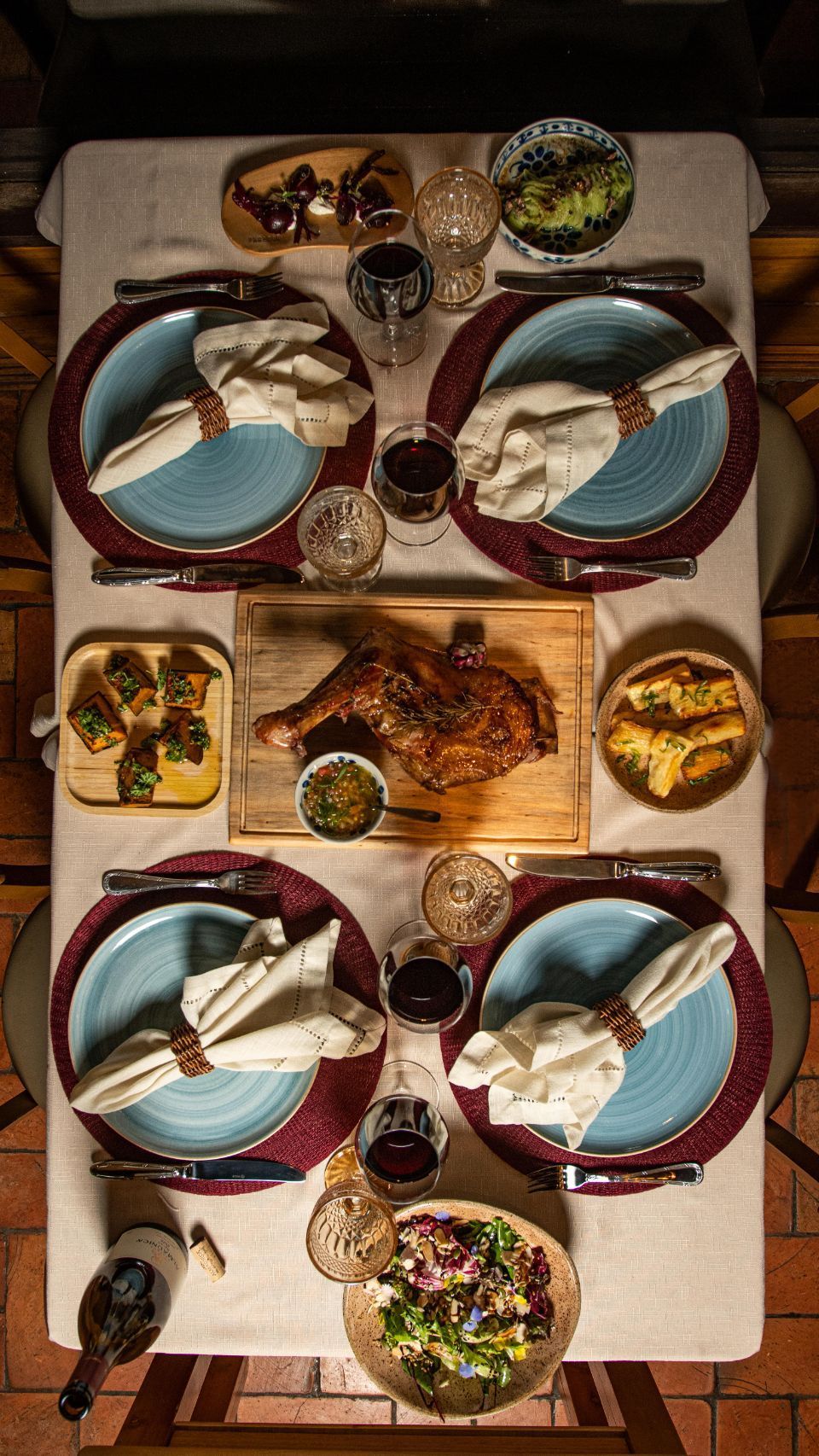 Mesa de jantar para quatro pessoas posta com pratos azuis, guardanapos brancos dobrados, tigelas de comida e bebidas sobre uma toalha de mesa branca.