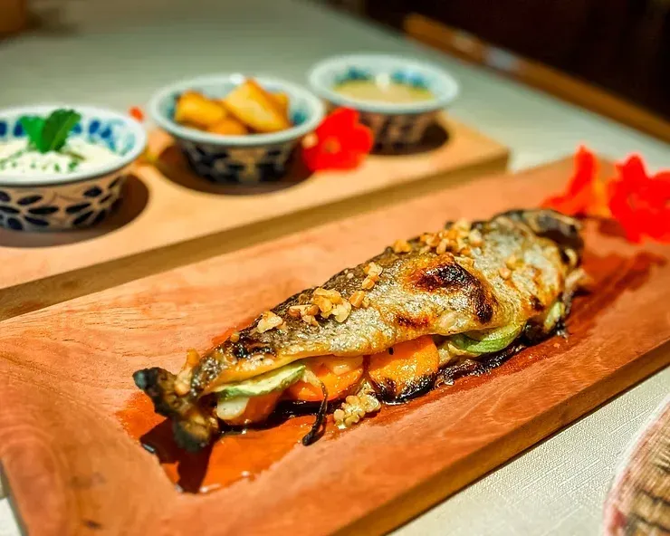 Peixe grelhado servido em uma tábua de madeira com acompanhamentos em uma mesa.