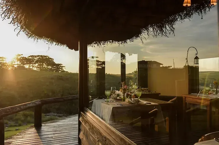Vista do pôr do sol num terraço de palha para refeições ao ar livre junto à água, com mesas e uma luz dourada e aconchegante.