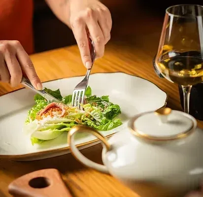 Mãos usando garfo e faca para comer salada em uma mesa com um bule de chá e uma taça de vinho.