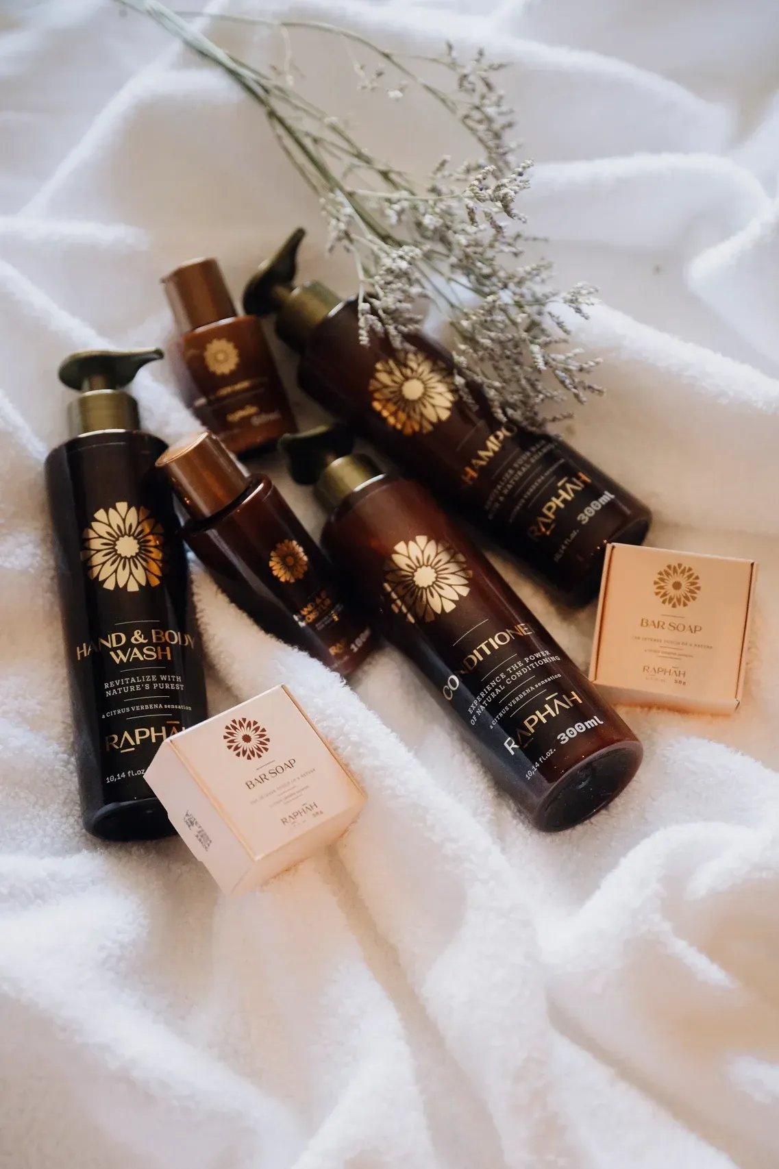 Diversos frascos e caixas de produtos de luxo para cuidados com a pele, dispostos sobre tecido branco com flores secas.