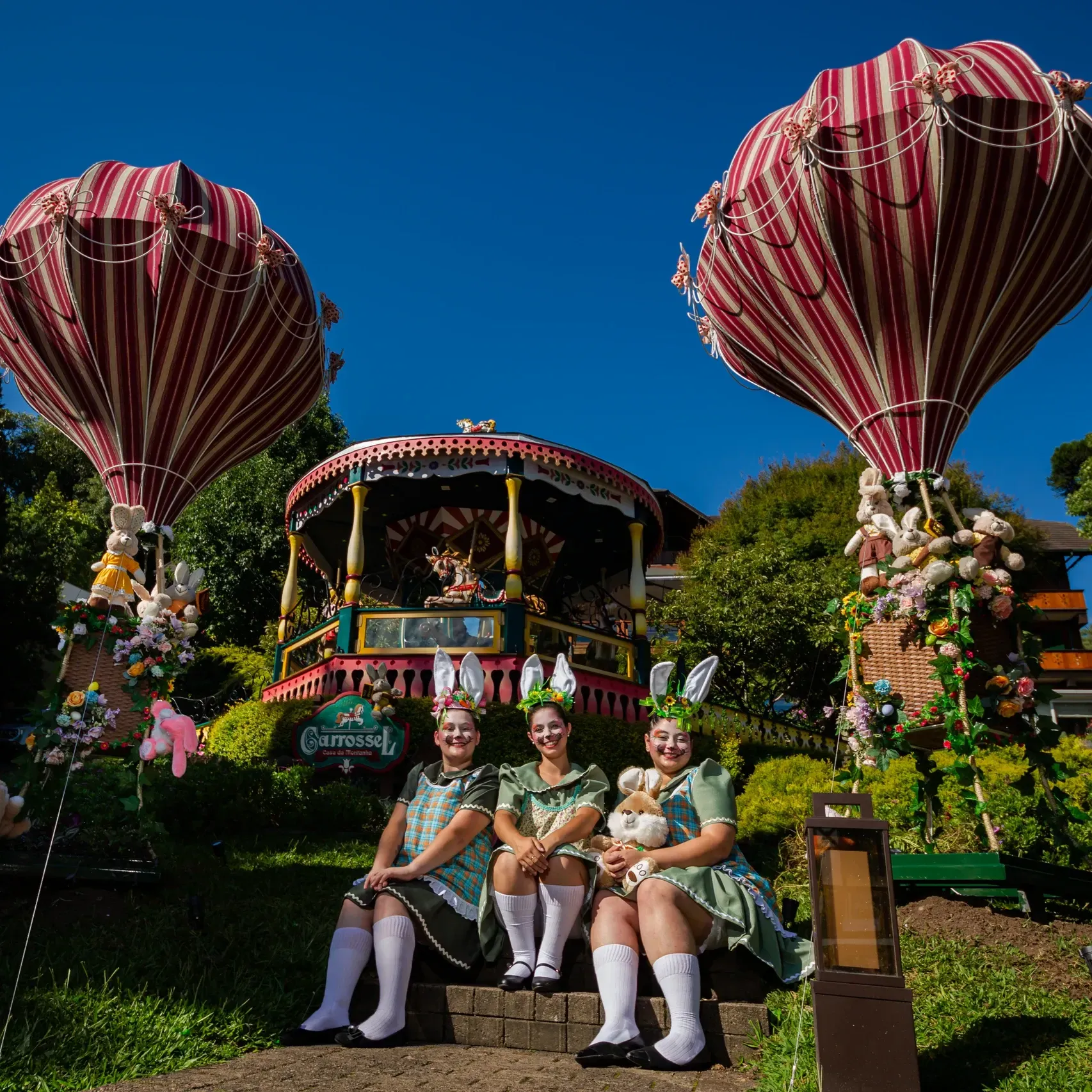 Três artistas vestidos de verde posam em frente a um carro alegórico decorado com grandes balões listrados sob um céu azul.