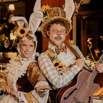 Artistas fantasiados com orelhas de coelho e girassóis, sorrindo e posando com um violão em um ambiente interno aconchegante.