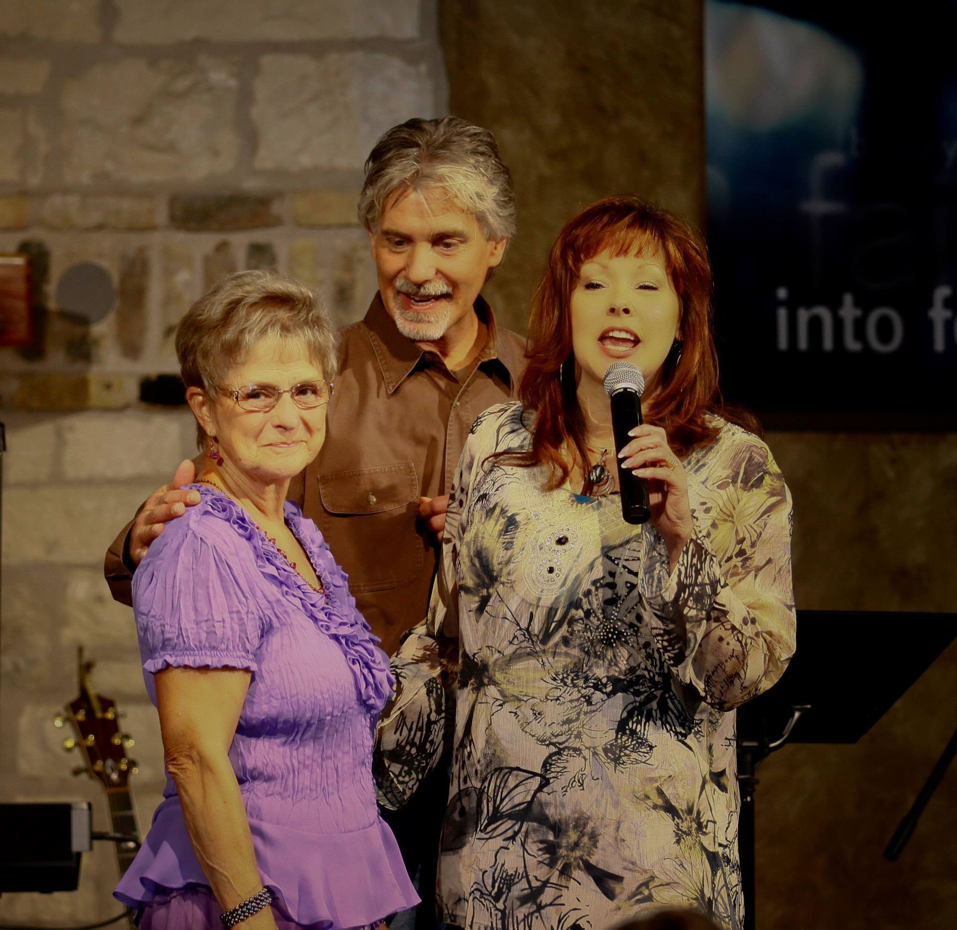 Three people stand on a stage with a stone background; one person sings into a microphone while others smile nearby.
