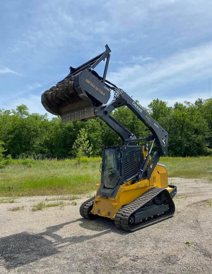 Yellow and black track skid steer with raised brush cutter attachment outdoors.