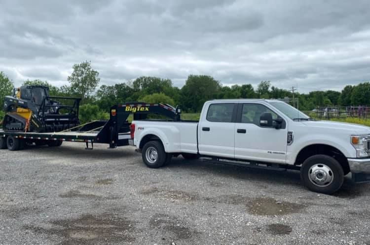 White pickup truck towing a trailer with a skid steer loader, on a gravel lot. Overcast sky.