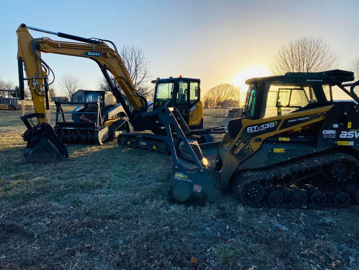 Construction equipment, including excavator and skid steer, on a grassy field at sunrise.