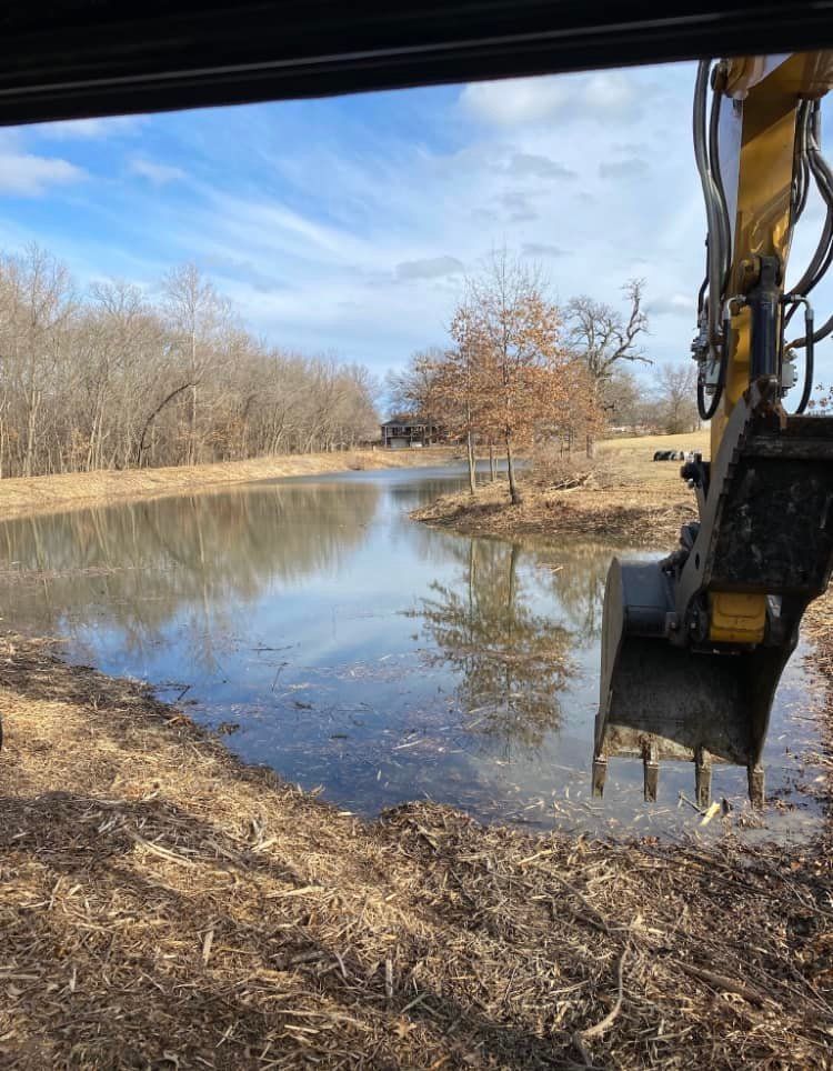 Excavator bucket near a pond reflecting trees under a blue sky.
