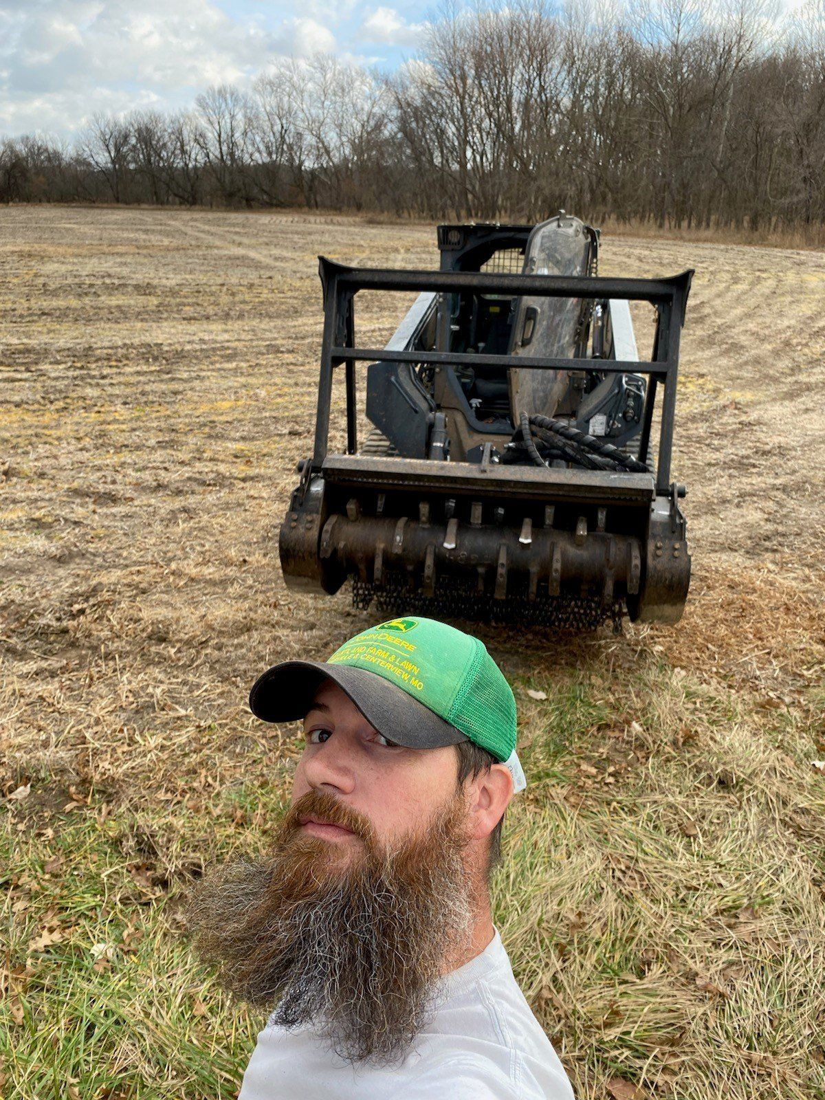 Man with long beard in front of a forestry mulcher in a field. He's wearing a green hat.