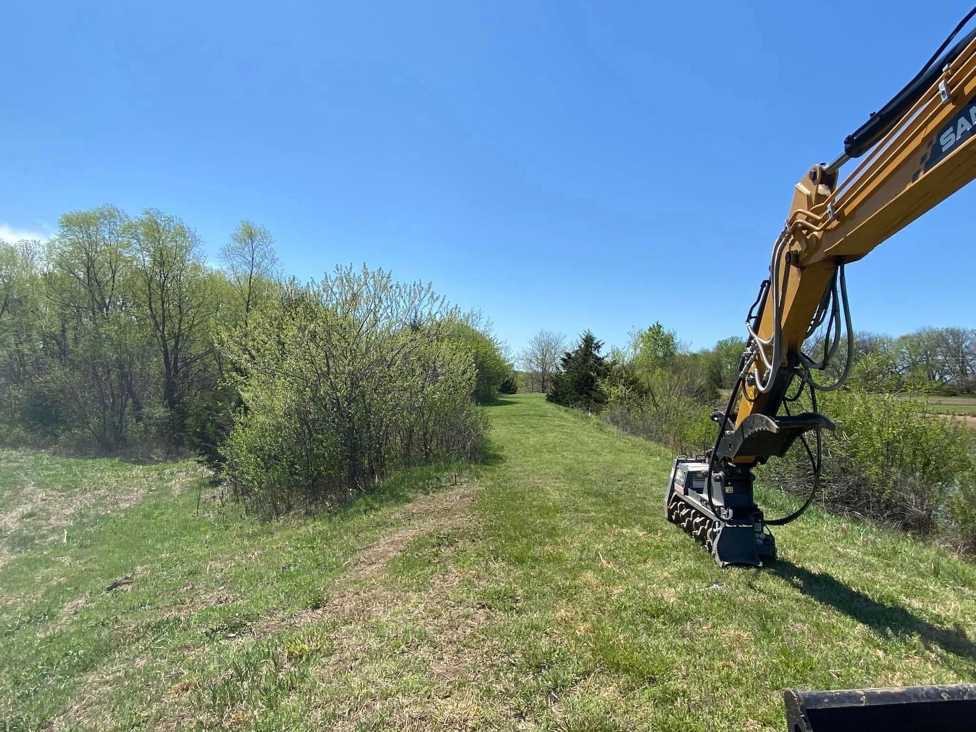 Excavator clearing a grassy path through trees and bushes on a sunny day.