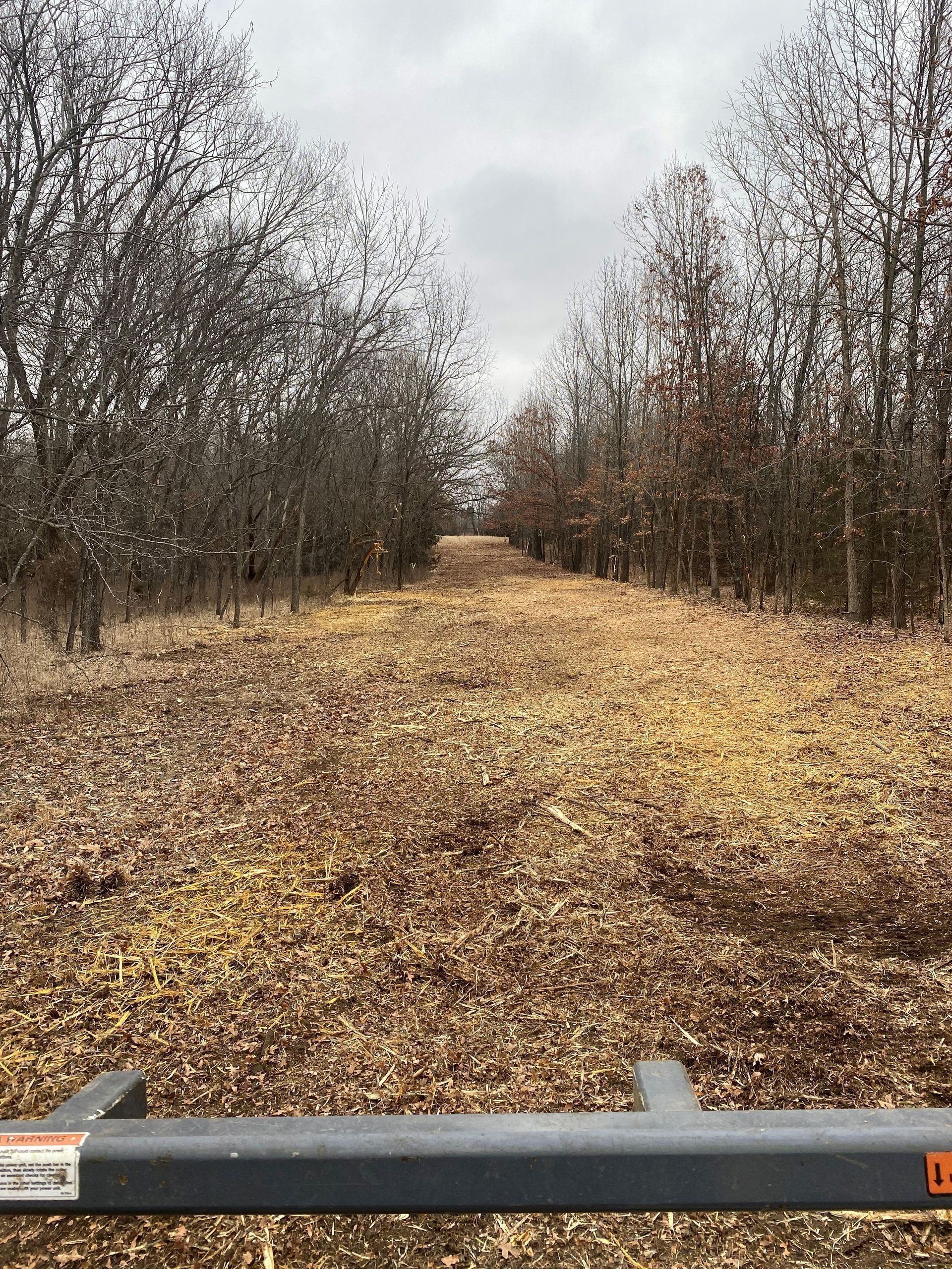 A leaf-covered path through a wooded area on a cloudy day. A gray tow bar is in the foreground.