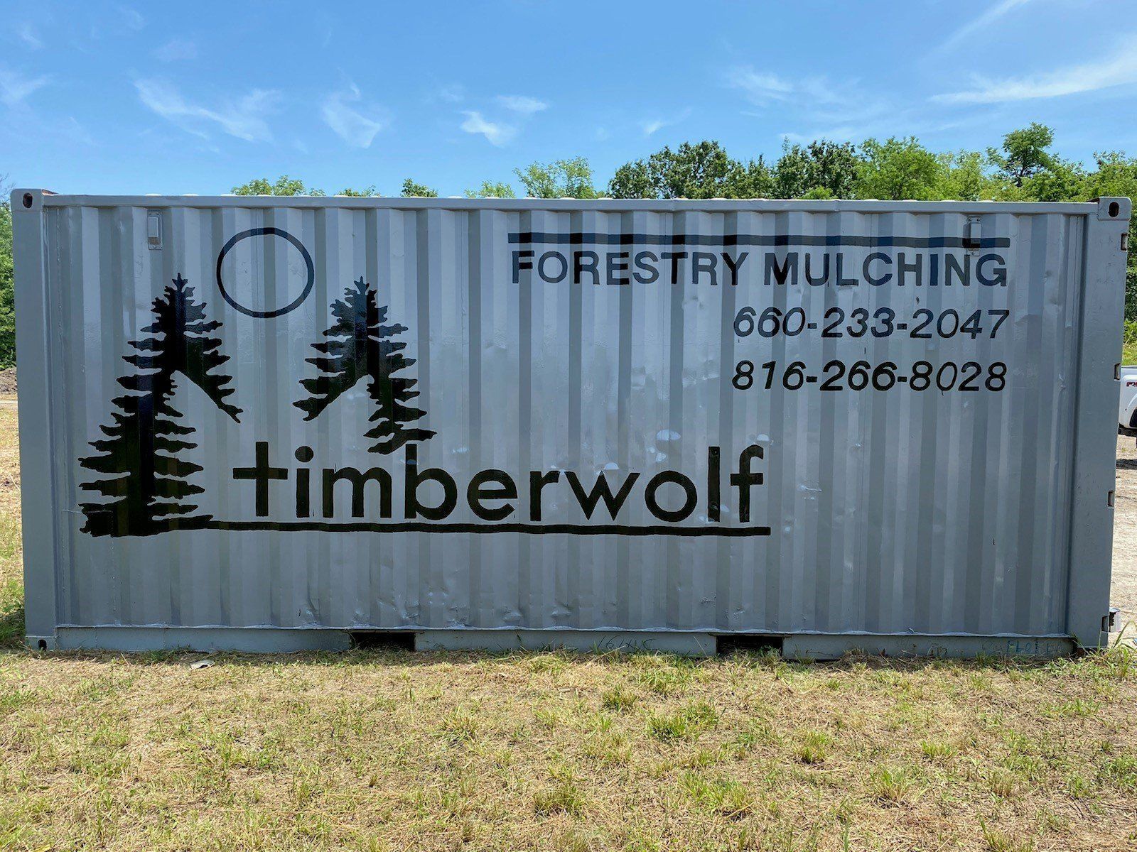 A silver shipping container with "timberwolf" logo, "Forestry Mulching" and phone numbers painted on the side.