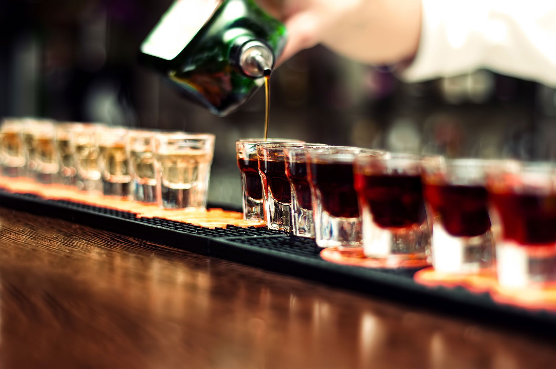 A bartender is pouring a drink into a row of shot glasses on a bar.
