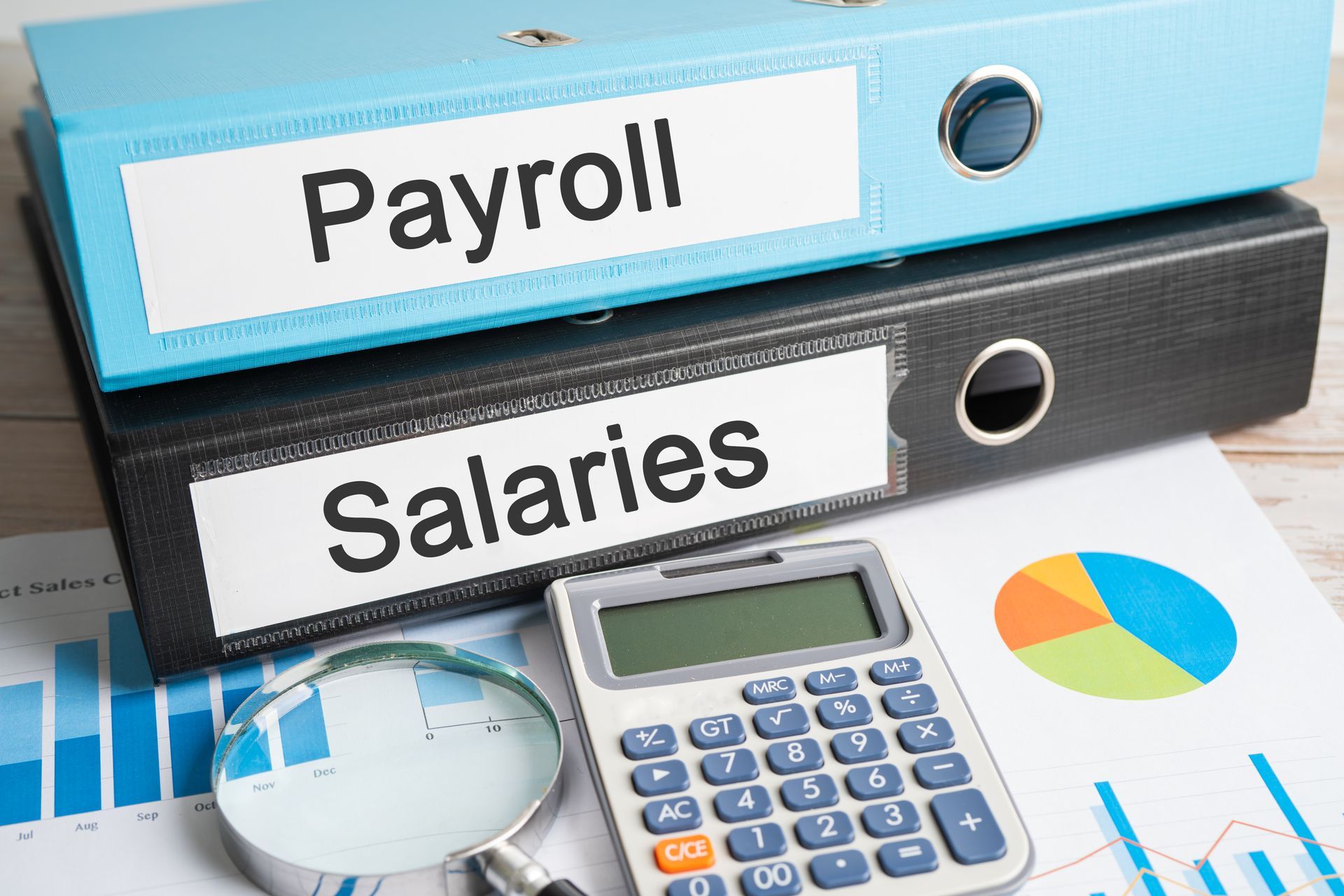 A binder labelled “payroll” and “salaries”. Documents with graphs, a calculator & a seeing eyeglass. A binder labelled “payroll” and “salaries”. Documents with graphs, a calculator & a seeing eyeglass.