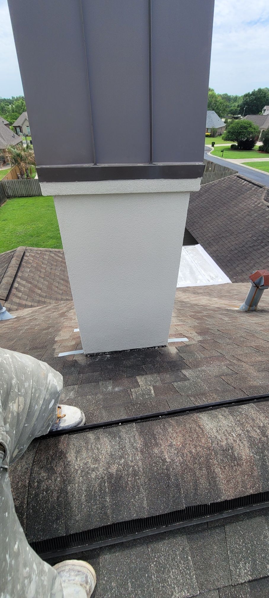 Gray rooftop chimney vent on shingles, with grassy yard and houses in the background.