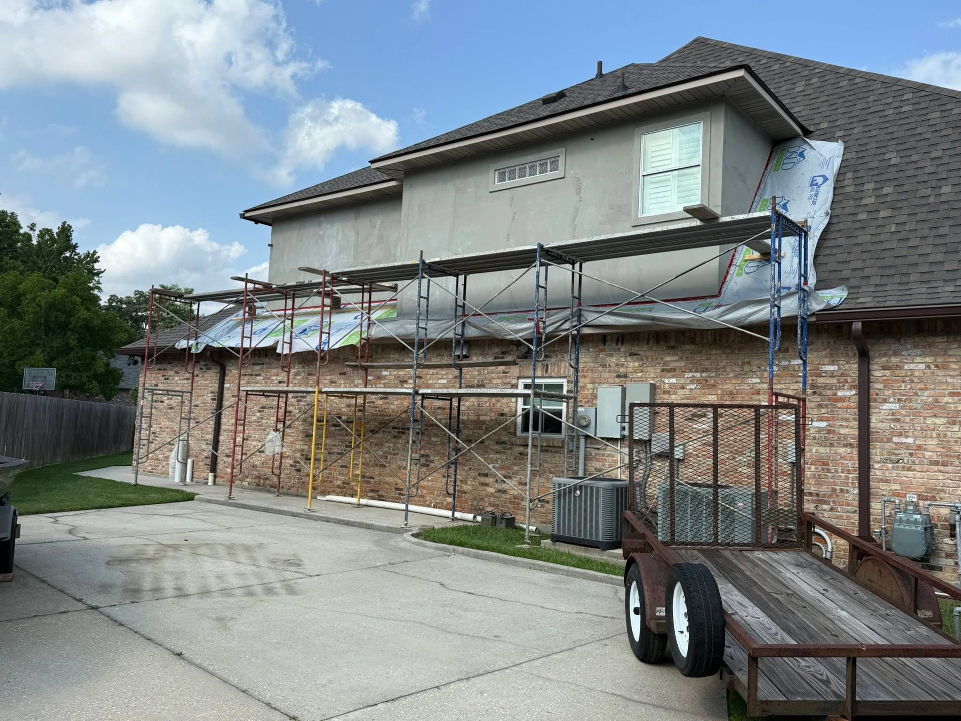 Scaffolding along brick house exterior with tarp-covered roof and utility trailer in driveway