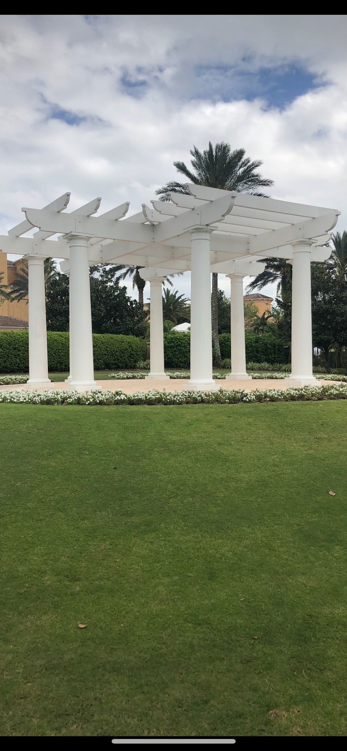 White pergola with leafy cutouts in a green lawn under a cloudy sky, with palm trees behind