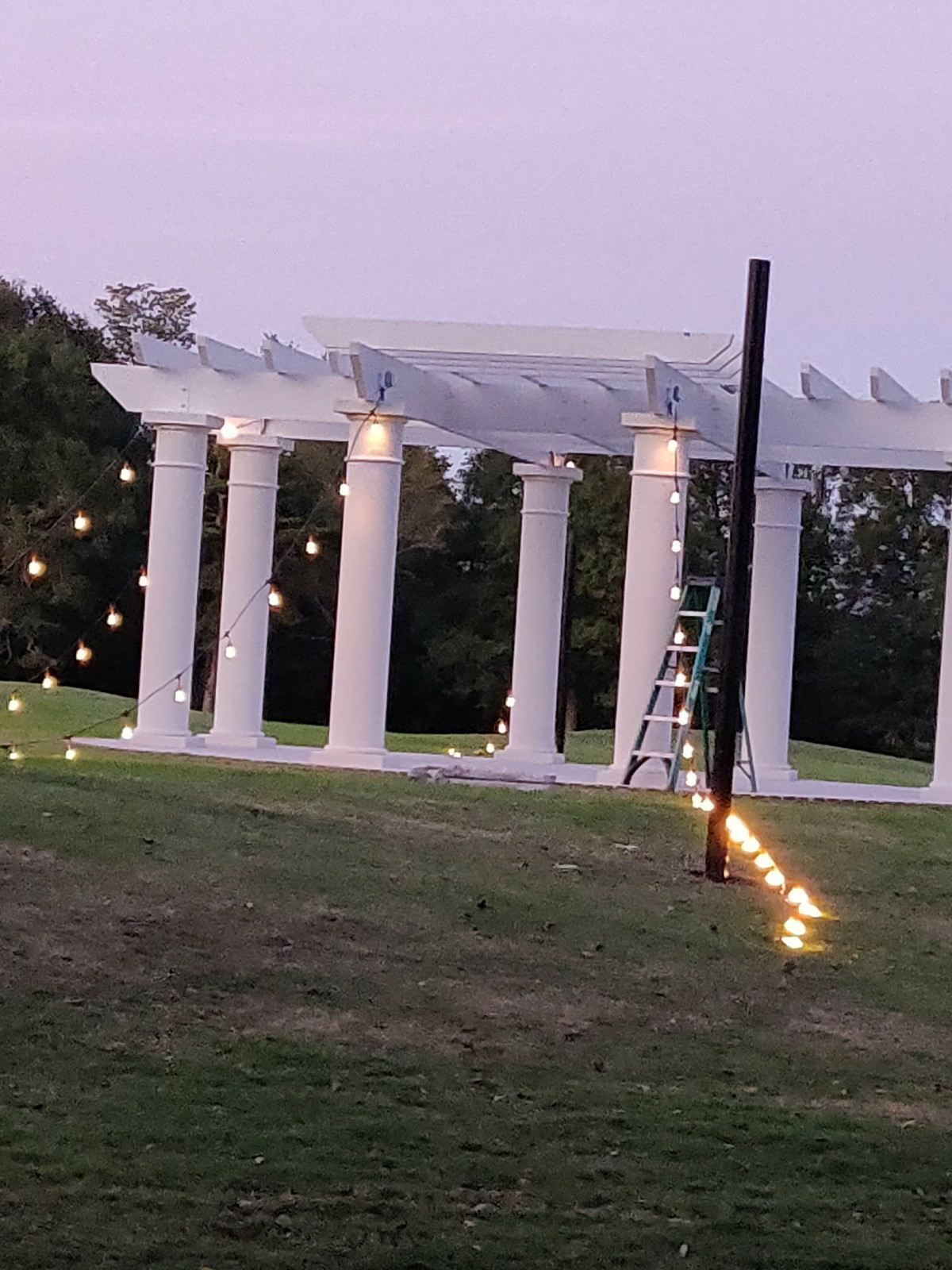 White pergola with columns at dusk, decorated with string lights on a grassy lawn, with a ladder nearby