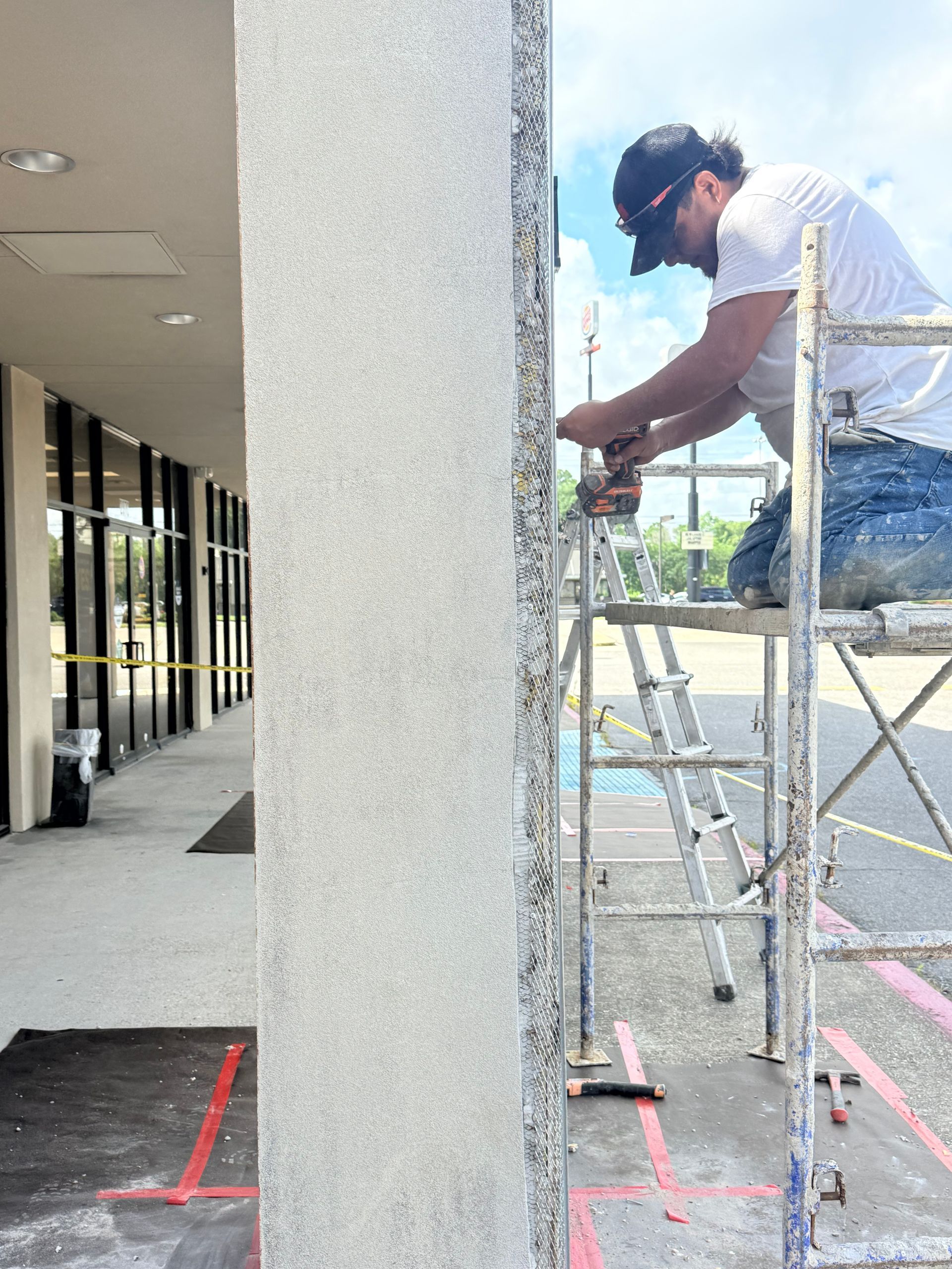 Worker on a ladder applying coating to an exterior wall beside a covered walkway.