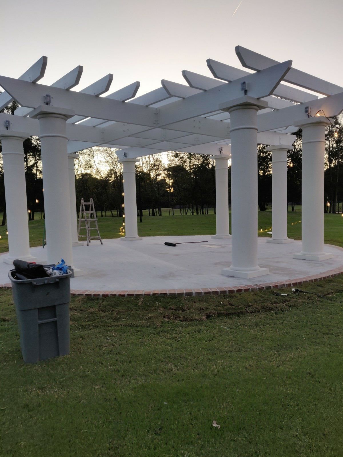 White pergola structure on a grassy lawn at dusk, with a trash can in the foreground and trees in back.