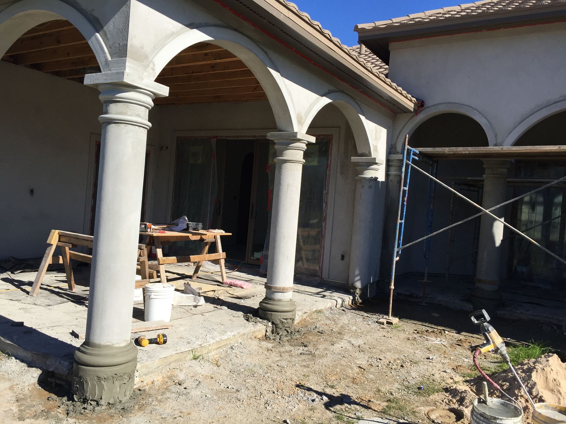House porch under construction with white columns, scaffolding, and tools on a dusty ground